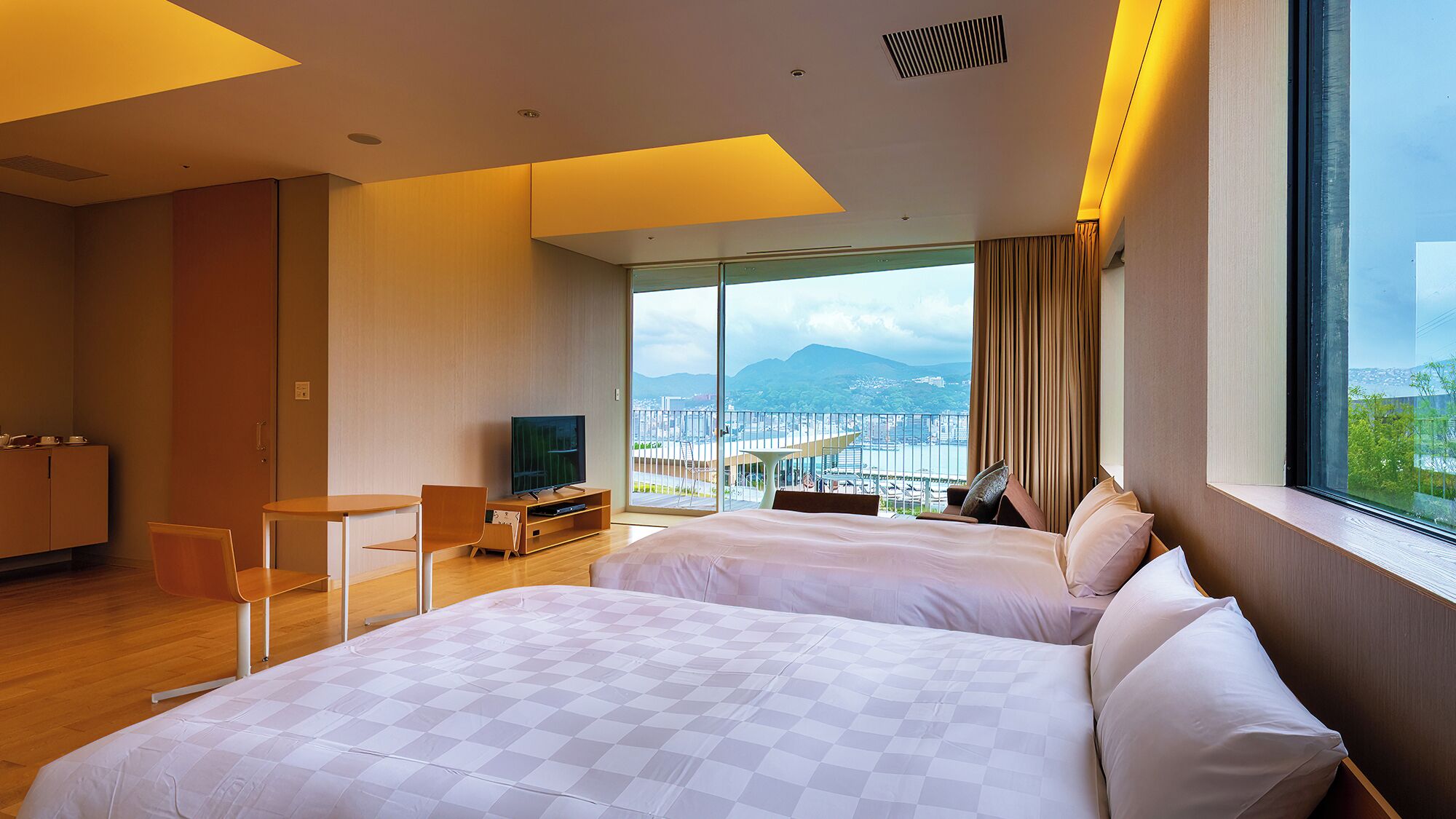 A hotel room with two beds, a desk, a TV, and a balcony with a city view.