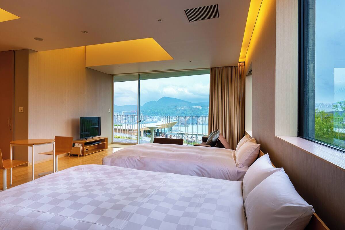 A hotel room with two beds, a desk, a TV, and a balcony with a city view.