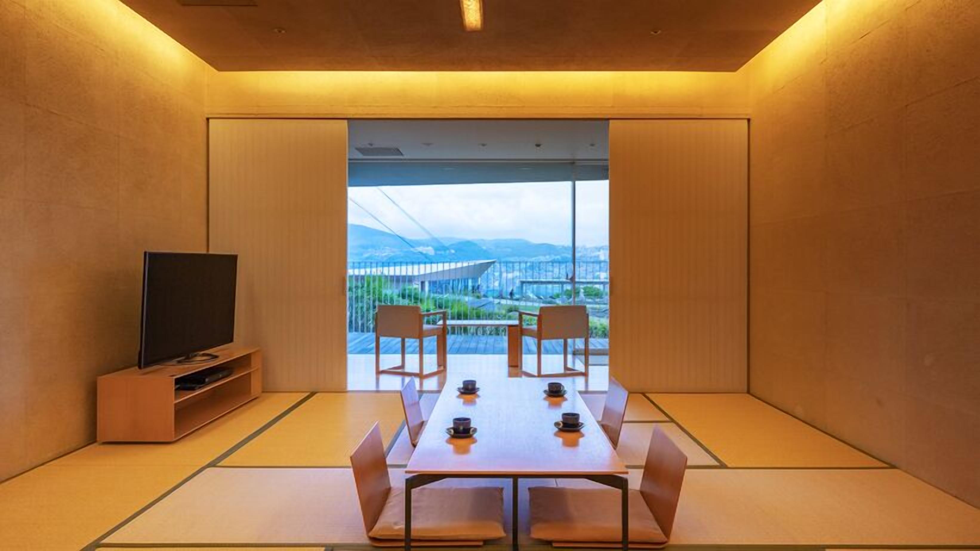 A room with tatami mats, a table with chairs, a television, and a view of a bridge and mountains.