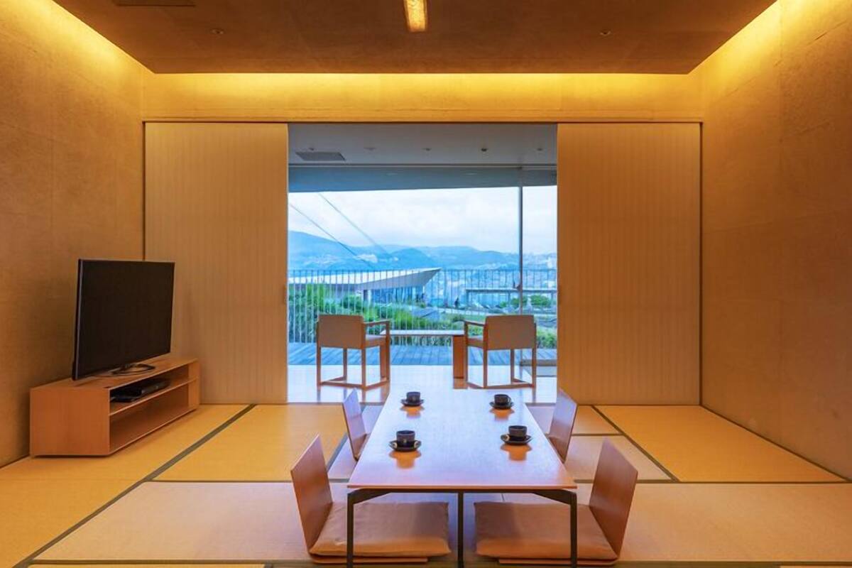 A room with tatami mats, a table with chairs, a television, and a view of a bridge and mountains.
