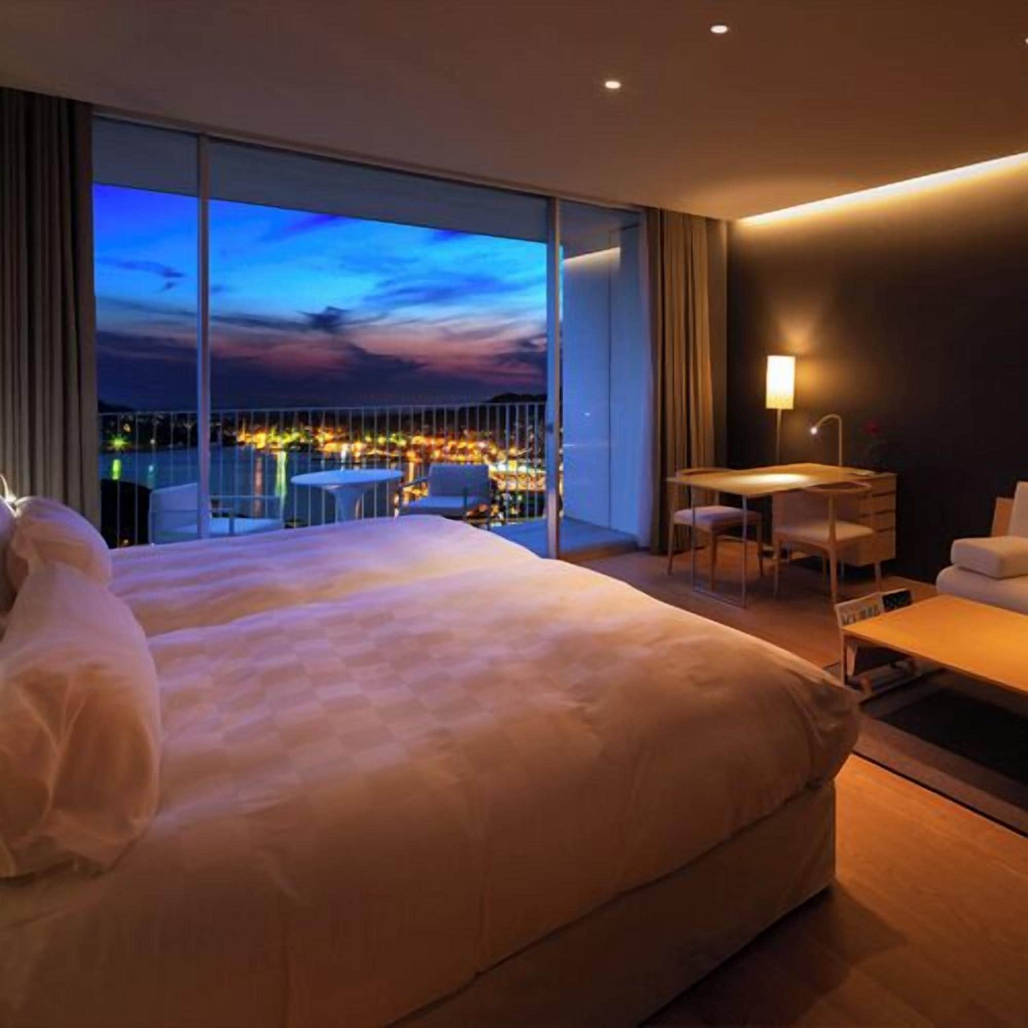 A hotel room with a large bed, a desk, and a chair, a view of the city at dusk, and a balcony with a table and chairs.