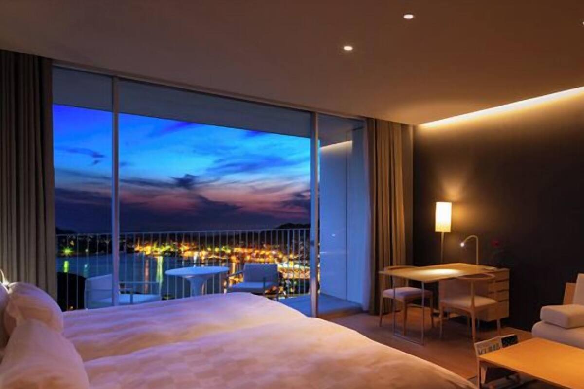 A hotel room with a large bed, a desk, and a chair, a view of the city at dusk, and a balcony with a table and chairs.