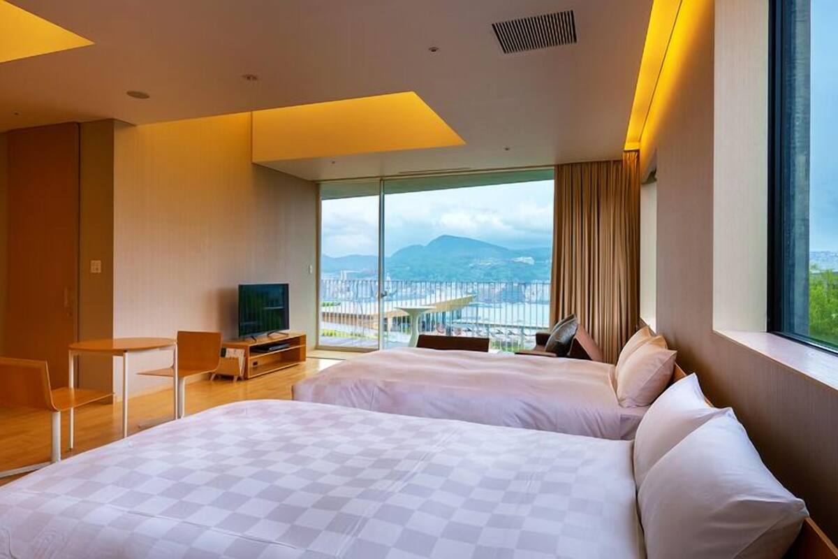 A hotel room with two beds, a TV, a desk, and a large window with a city view.