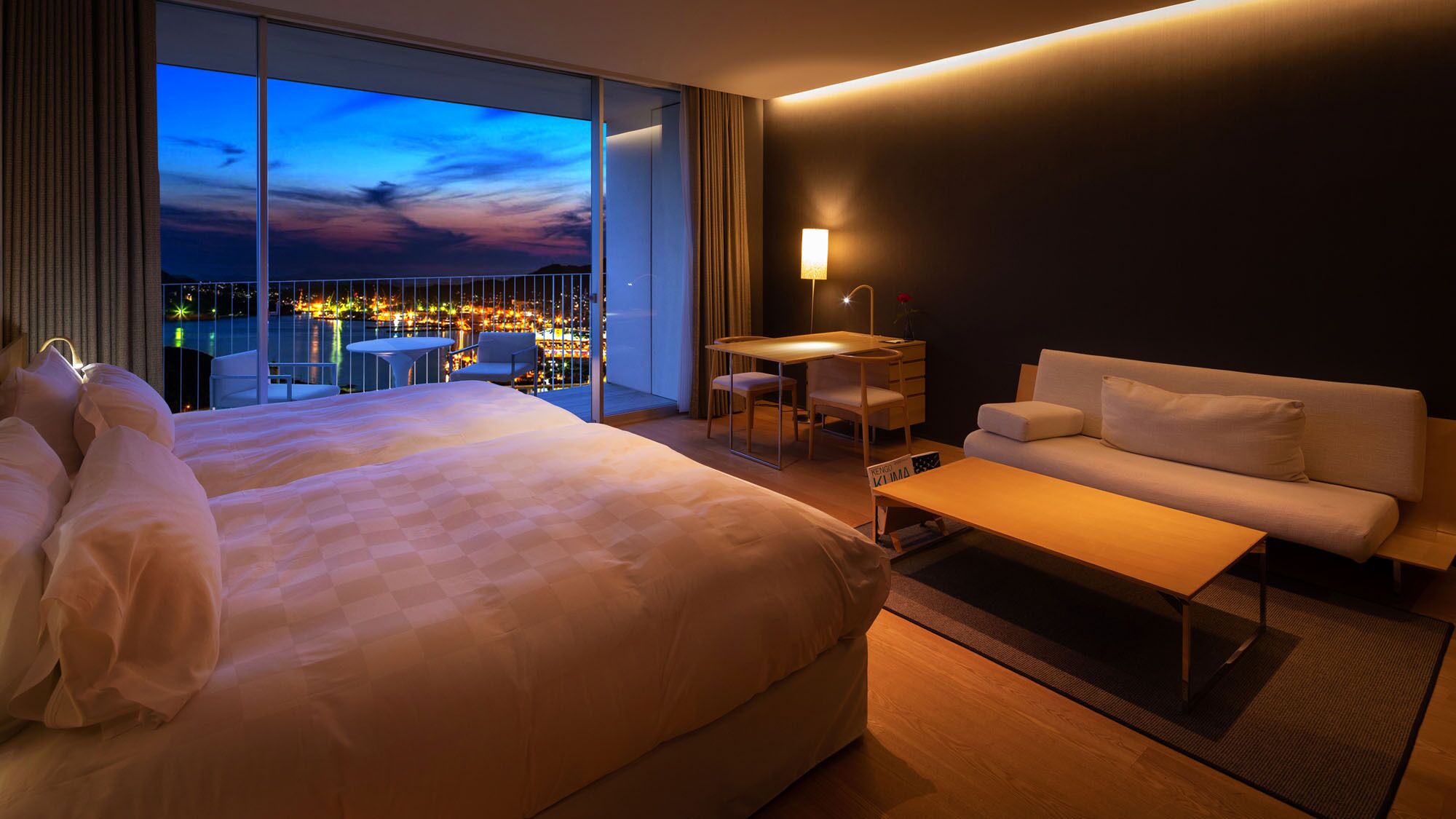 A hotel room with a large bed, a sofa, a desk, and a view of a cityscape at dusk.
