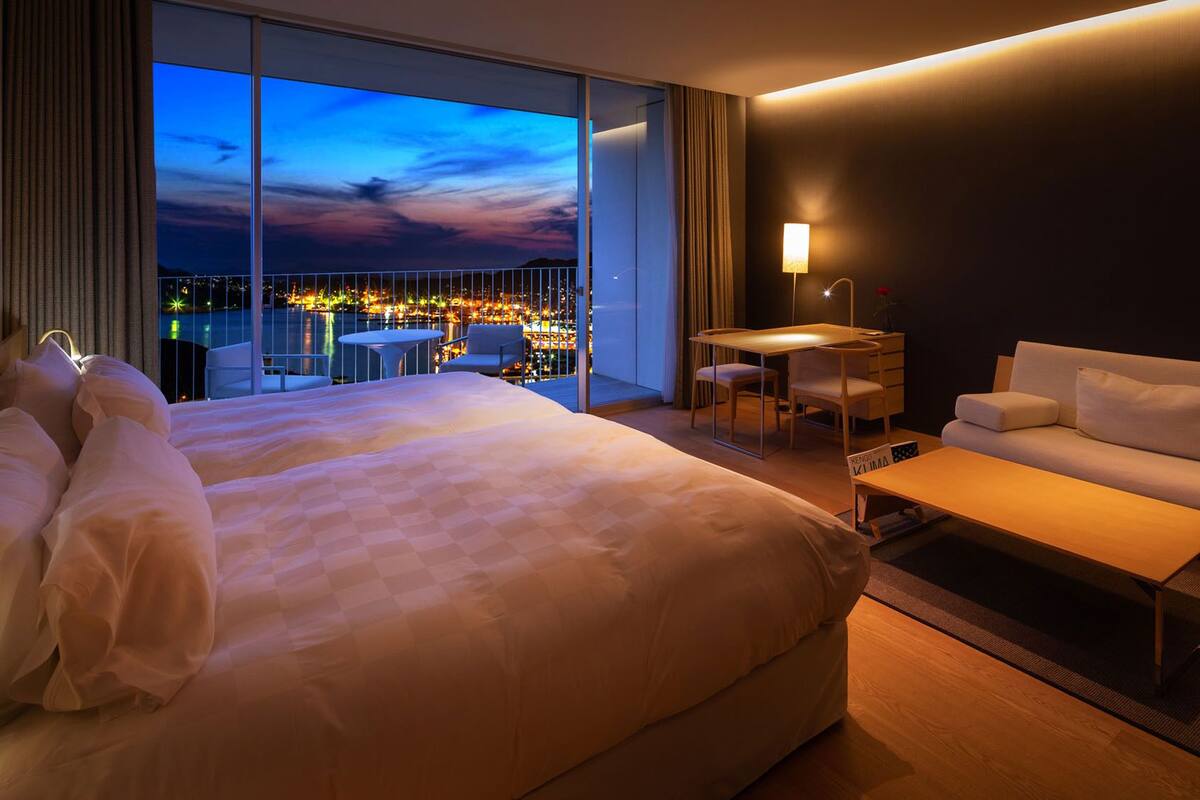 A hotel room with a large bed, a sofa, a desk, and a view of a cityscape at dusk.