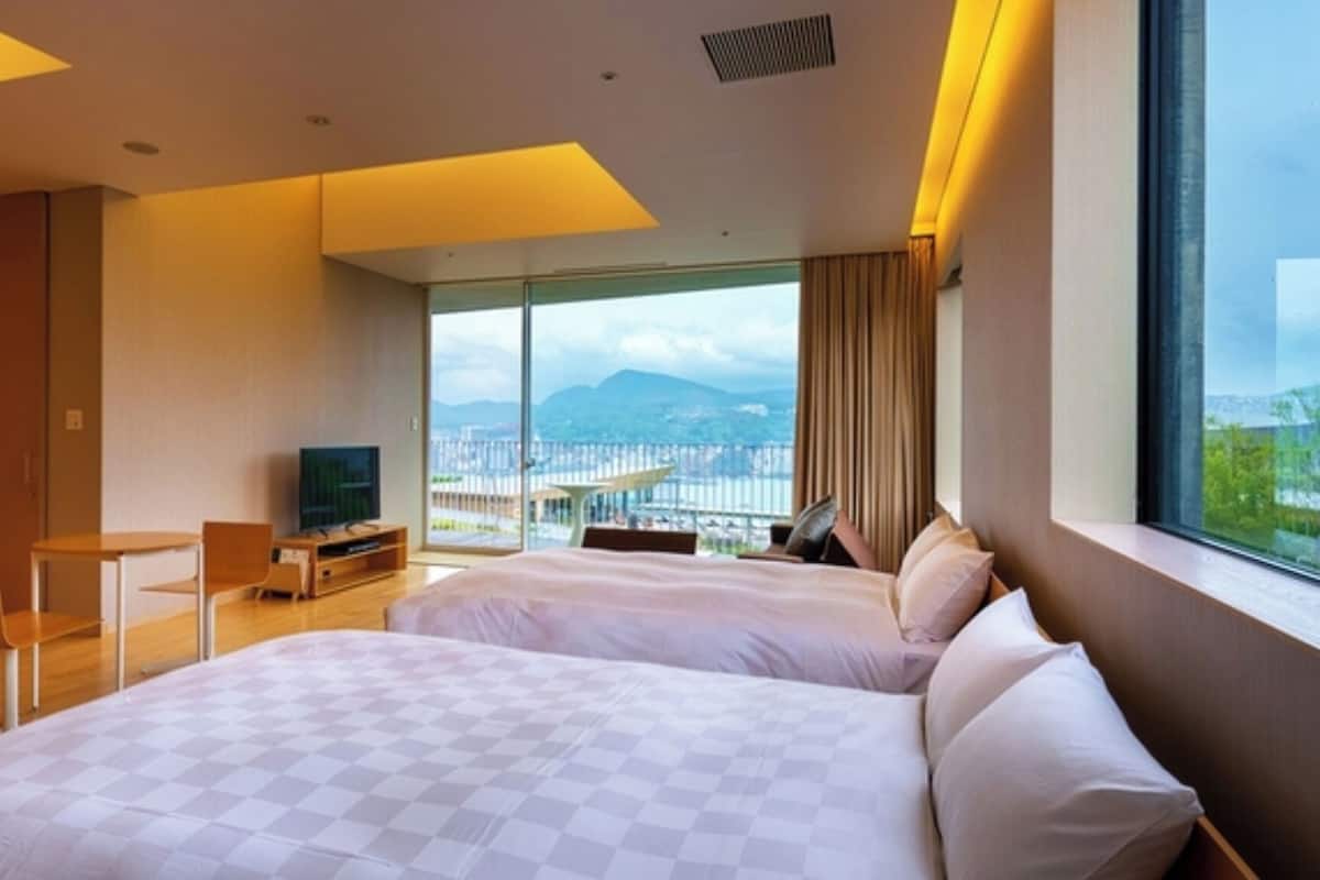 A modern hotel room with two beds, a TV, a desk, and a large window with a city view.