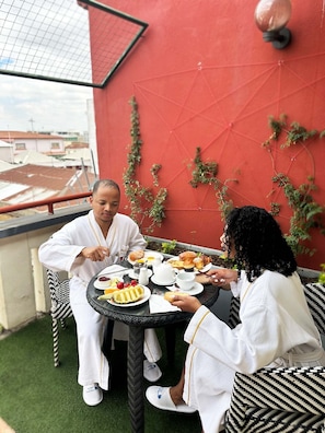 A dining table with plates of food, a red wall with climbing plants, and a view of buildings in the background. - Le Grand Mellis Hôtel & Spa (Antananarivo)
