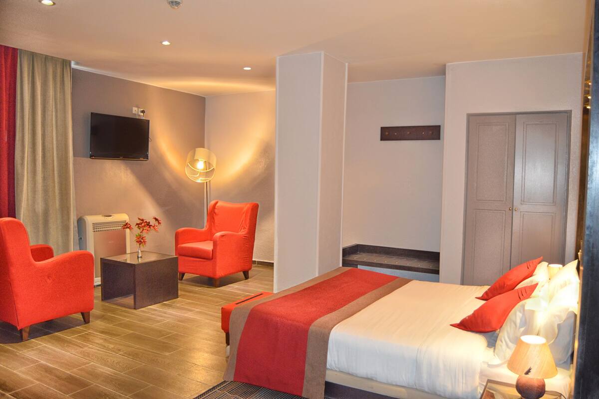 A hotel room with a bed, two red armchairs, a small table with a vase of flowers, a television mounted on the wall, and a door with a handle.