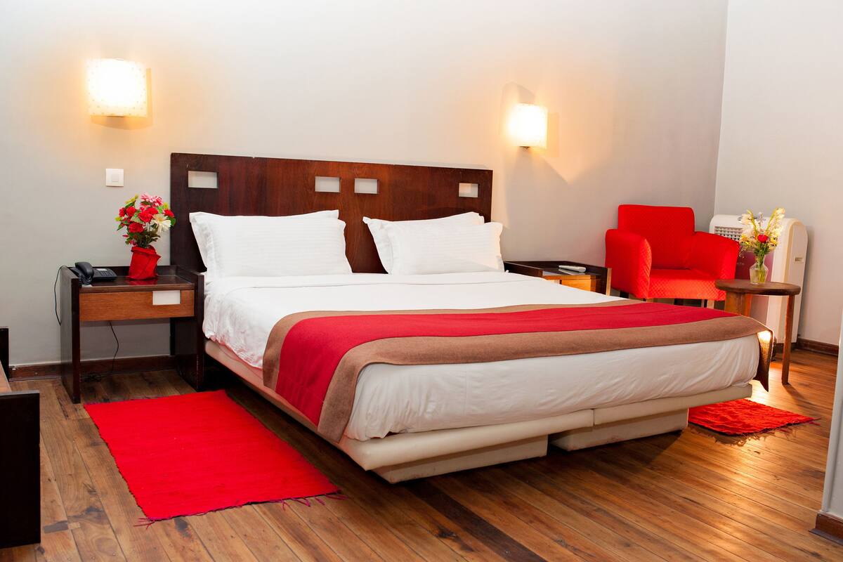 A hotel room with a large bed, two bedside tables, a red chair, a small table with a vase of flowers, and a red rug.