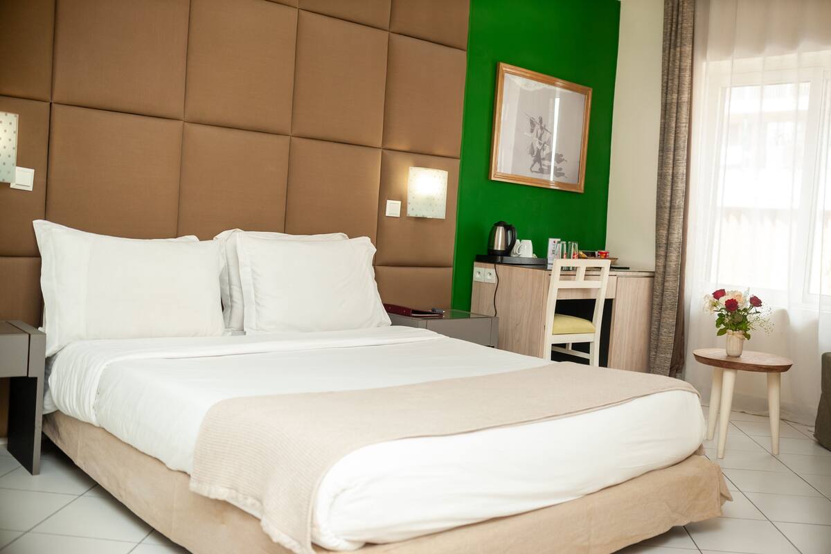 A hotel room with a large bed, a desk, a chair, a small table with a vase of flowers, and a green accent wall.