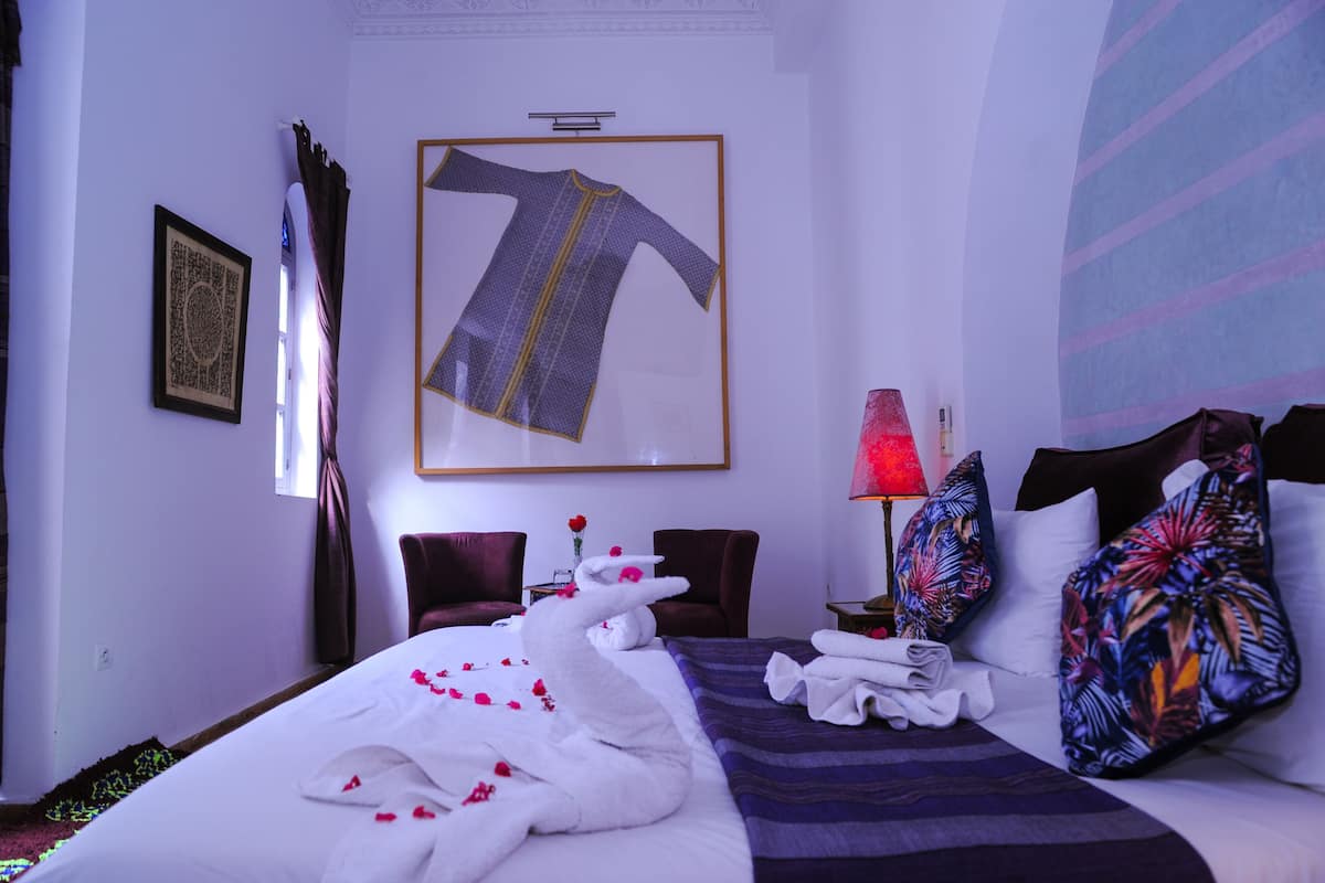 A hotel room with a neatly made bed, a framed garment on the wall, a red lamp, and tropical patterned pillows.