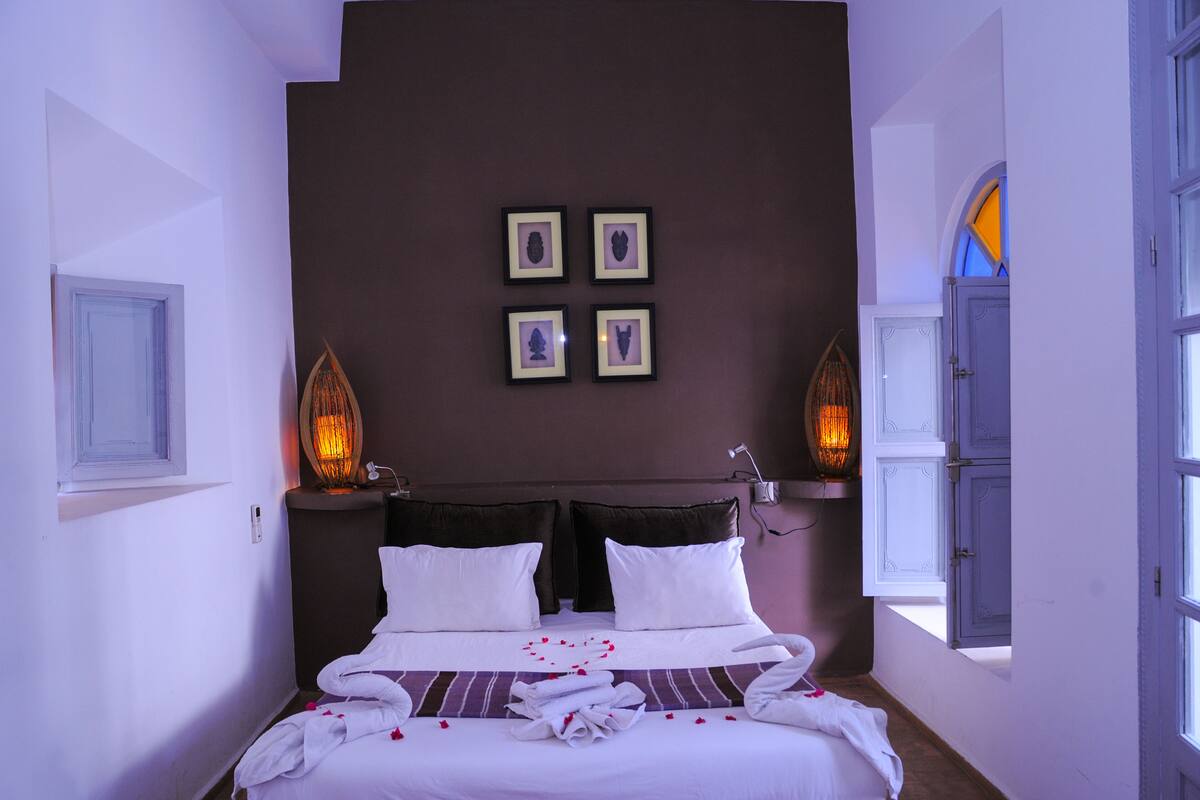 A neatly made bed with white linens and a heart-shaped arrangement of rose petals. There are four framed pictures on the wall above the bed.