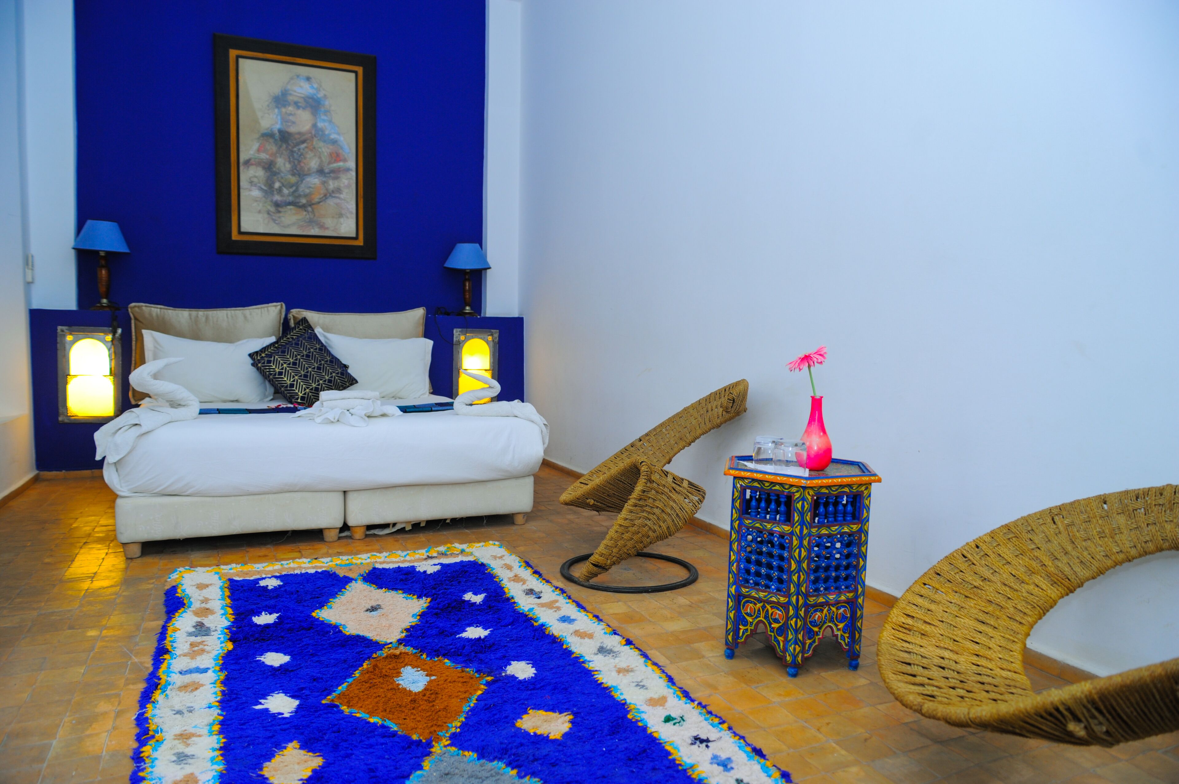 Une chambre à coucher avec un lit, un mur bleu, un tableau, une table de chevet avec une lampe, un vase et une chaise en osier.