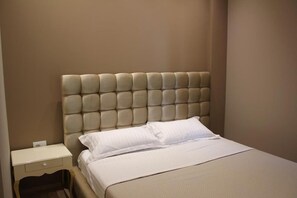 Quarto com cabeceira estofada, uma cama de solteiro com roupa de cama branca e mesa de cabeceira com gaveta.