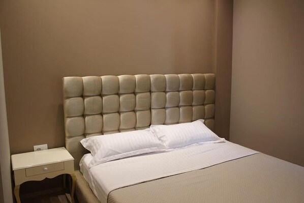 Quarto com cabeceira estofada, uma cama de solteiro com roupa de cama branca e mesa de cabeceira com gaveta.