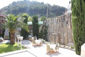 Um pátio externo com mesas e cadeiras, cercado por vegetação e um muro de pedra.