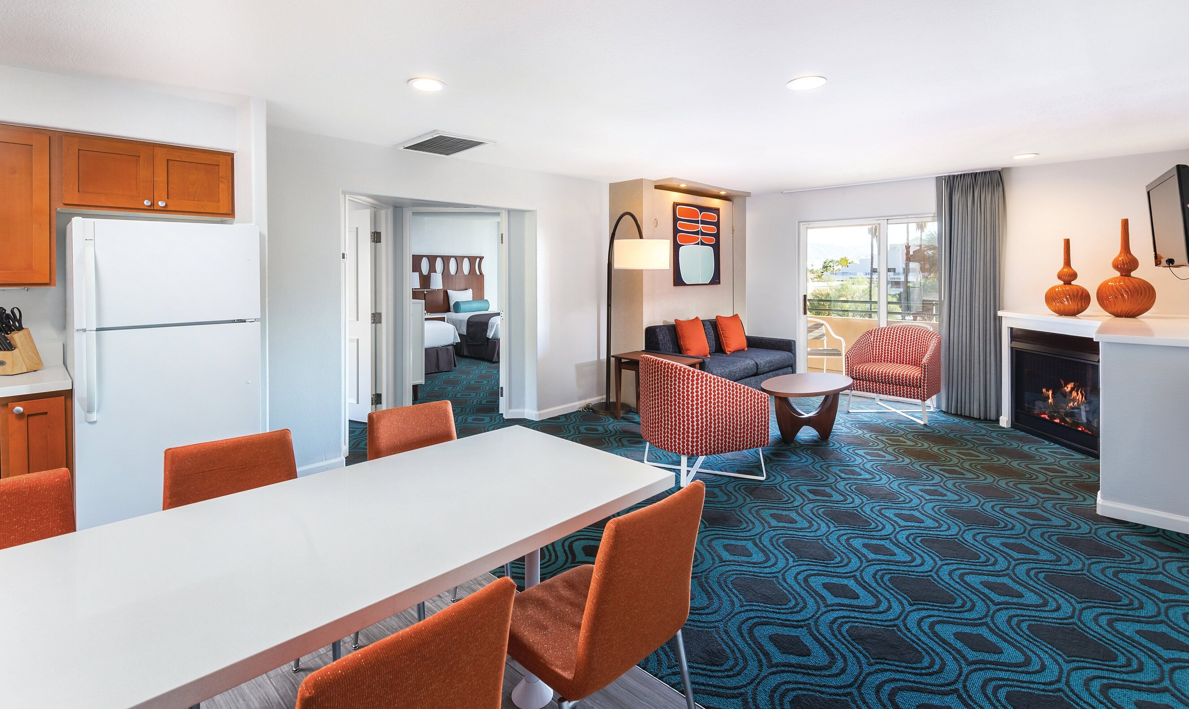 A modern hotel room with a dining area, a living room with a sofa and armchairs, a fireplace, and a kitchen with wooden cabinets and a refrigerator.