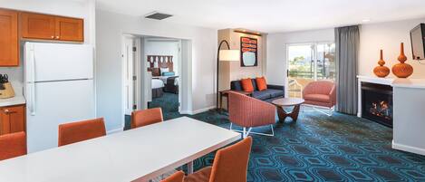 A modern hotel room with a dining area, a living room with a sofa and armchairs, a fireplace, and a kitchen with wooden cabinets and a refrigerator.