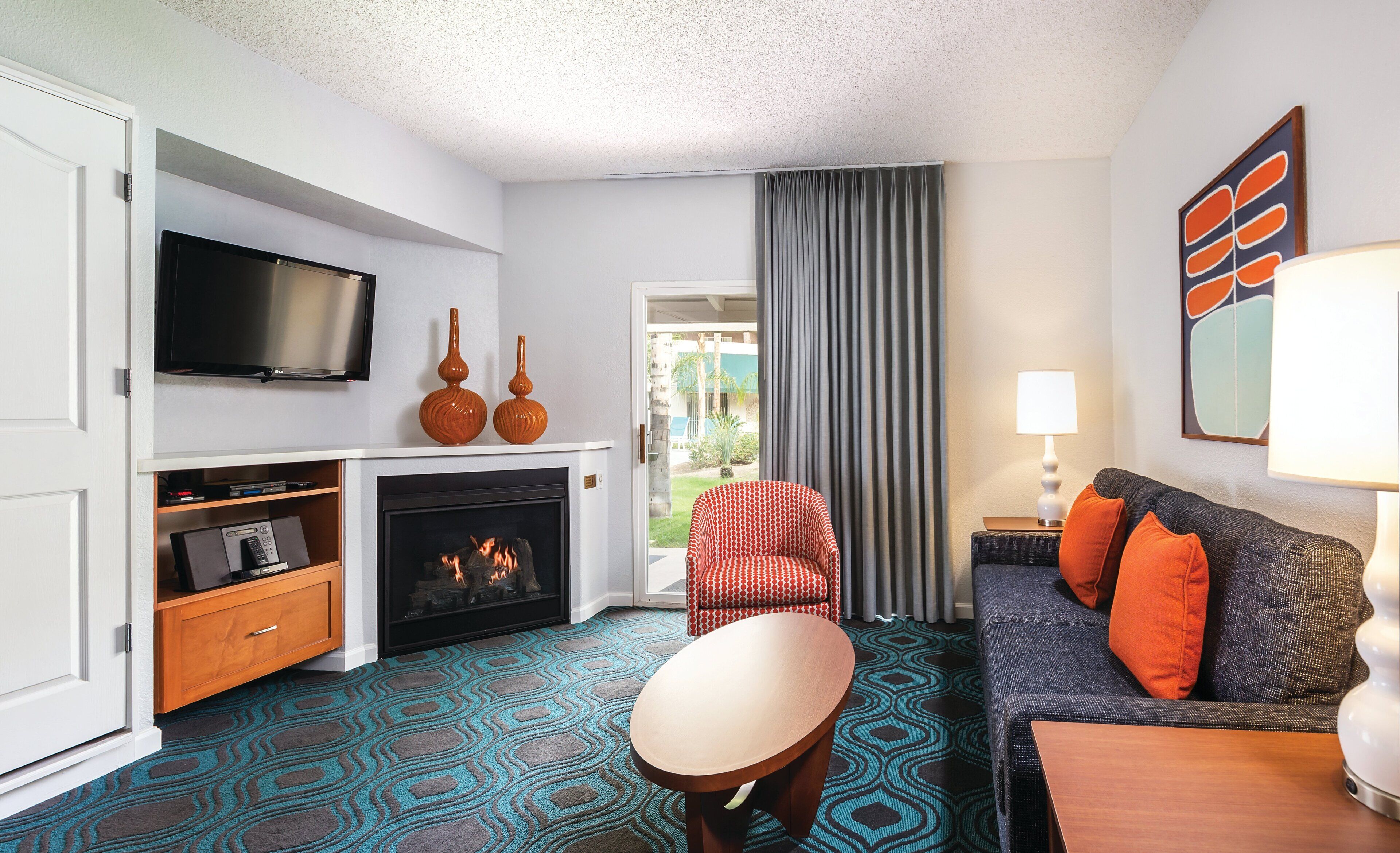 A modern hotel room with a flat-screen TV, a fireplace, a sofa with orange cushions, a patterned carpet, and a view of the outdoors.