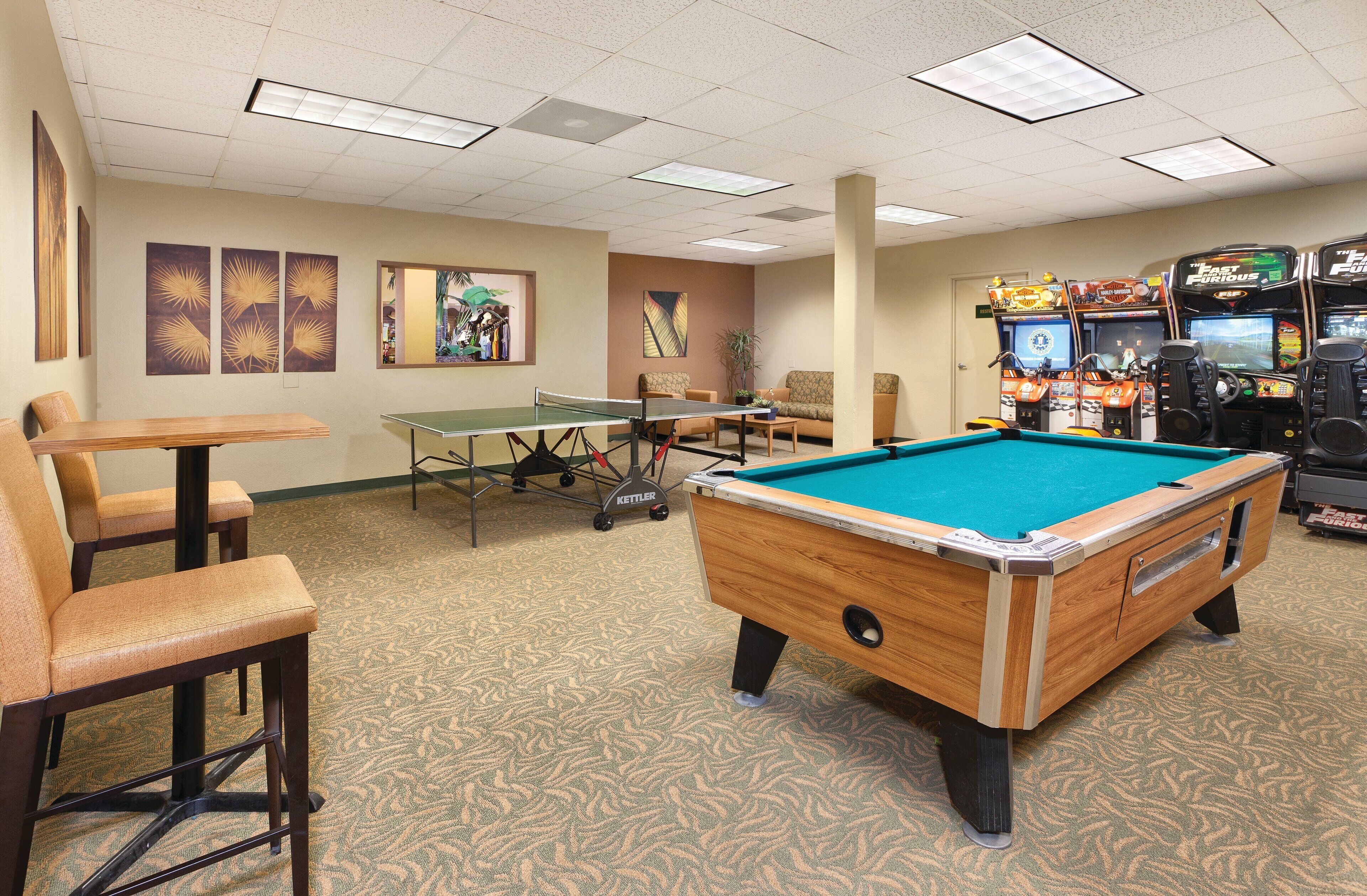 Une table de billard, une table de ping-pong et des jeux d’arcade dans une pièce dotée d’un tapis à motifs et d’œuvres d’art mural.