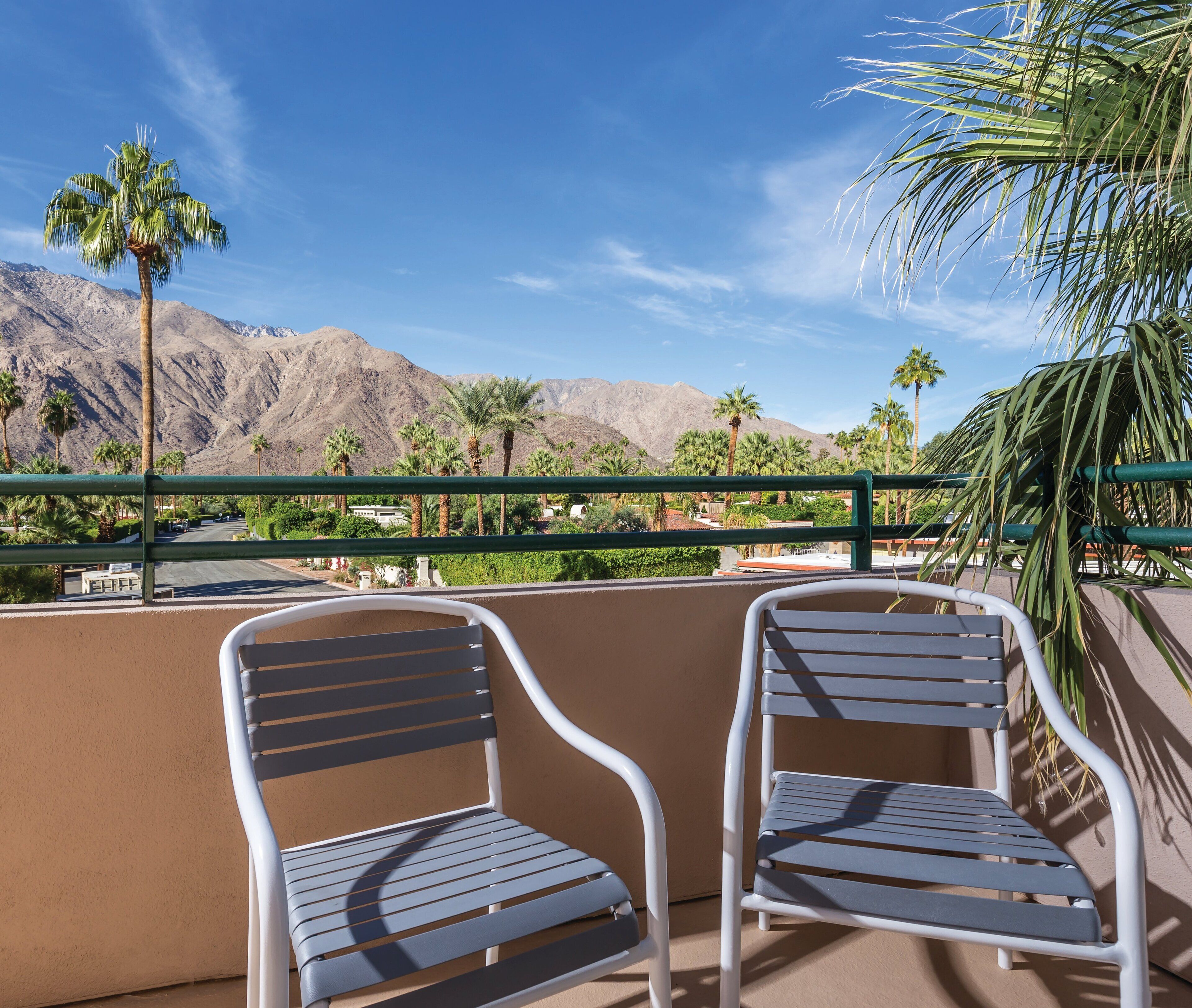 Two metal chairs on a balcony overlooking a mountainous landscape with palm trees.