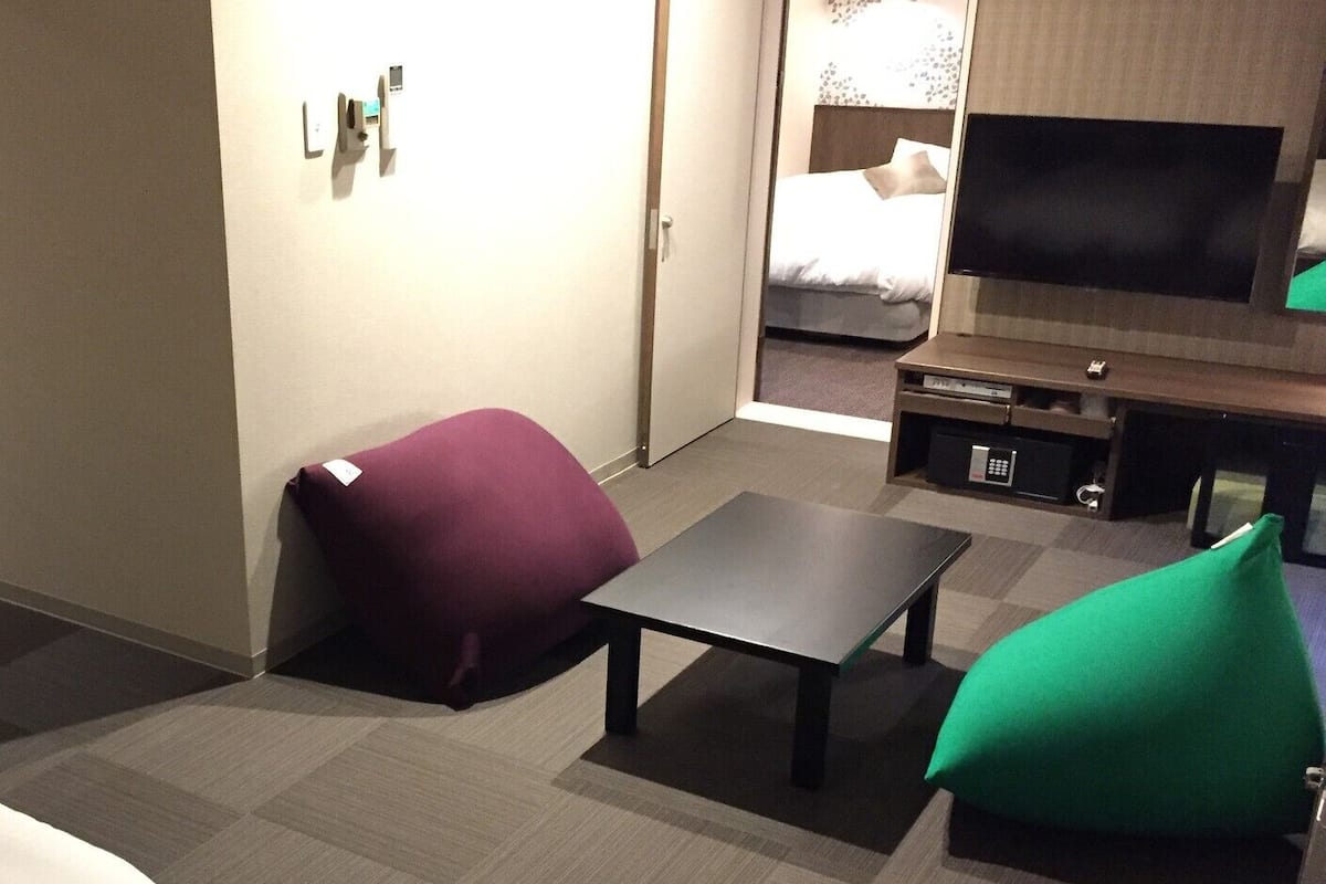 Habitación de hotel con cama, televisor, una mesita y dos pufs.