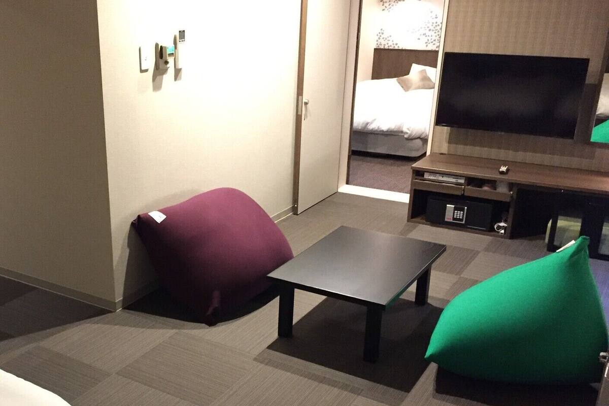 Habitación de hotel con cama, televisor, una mesita y dos pufs.