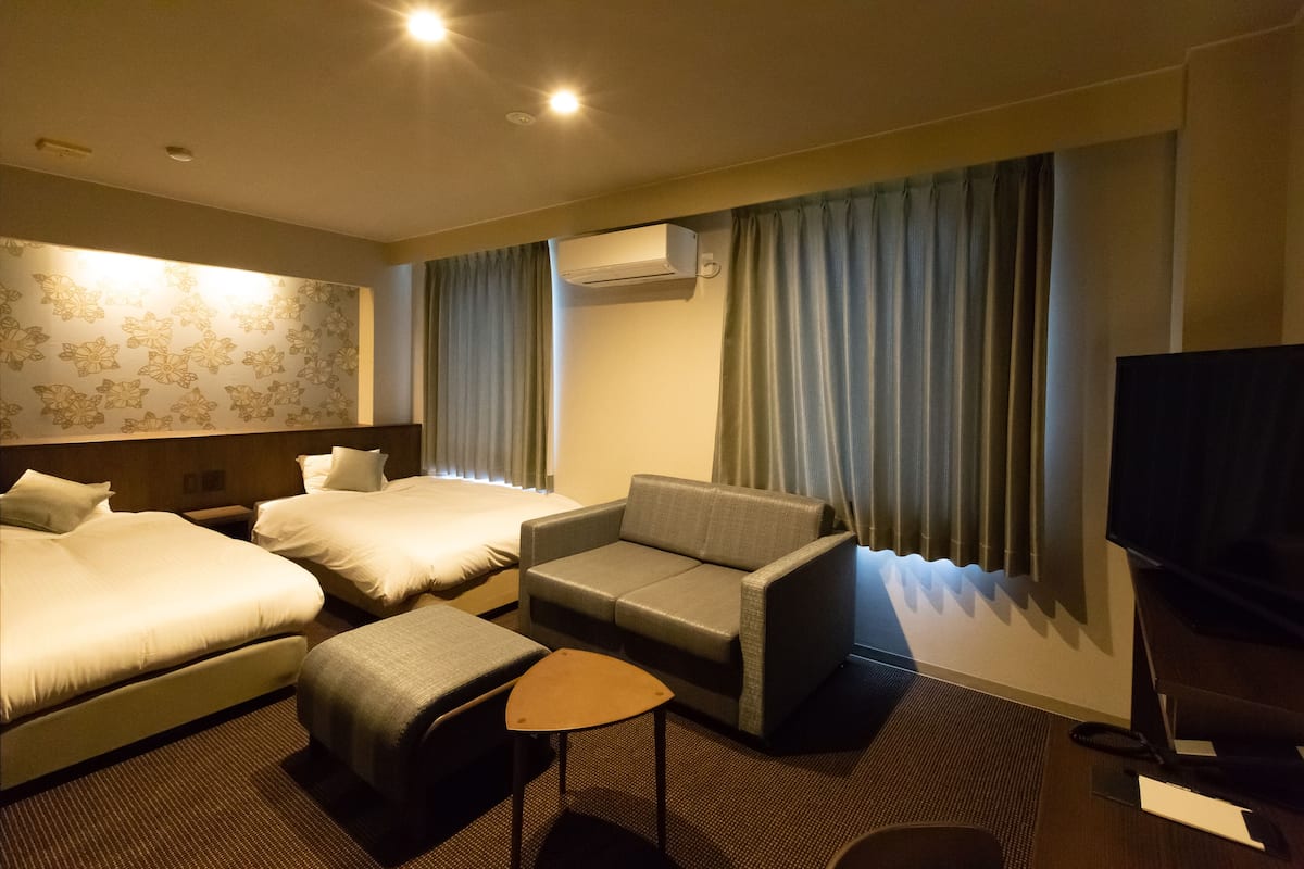 Habitación de hotel con dos camas, un sofá, una mesita pequeña, aire acondicionado y televisor.