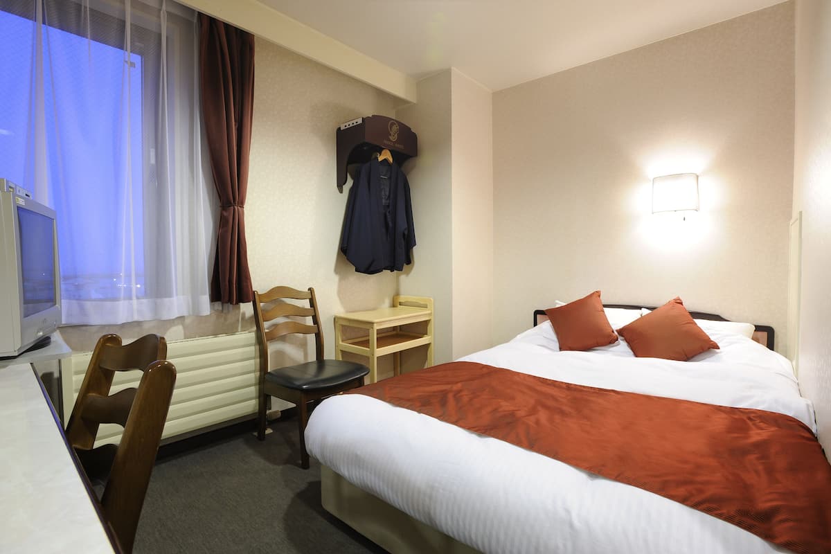 Habitación de hotel con cama, escritorio, silla, perchero y ventana con cortinas.