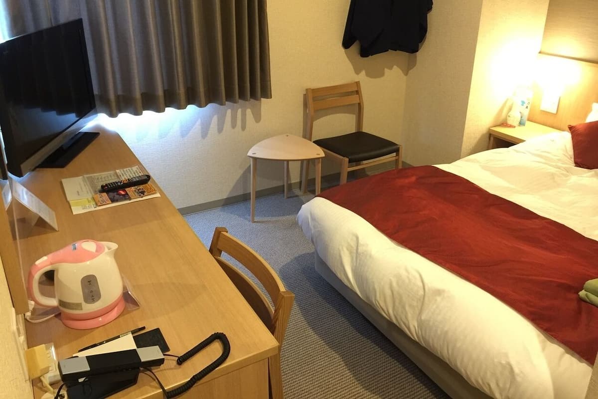 Una habitación de hotel con una cama, un escritorio con hervidor, una silla, un televisor y una lámpara.