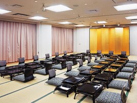 A conference room with rows of tables and chairs, large windows with curtains, and a stage area.