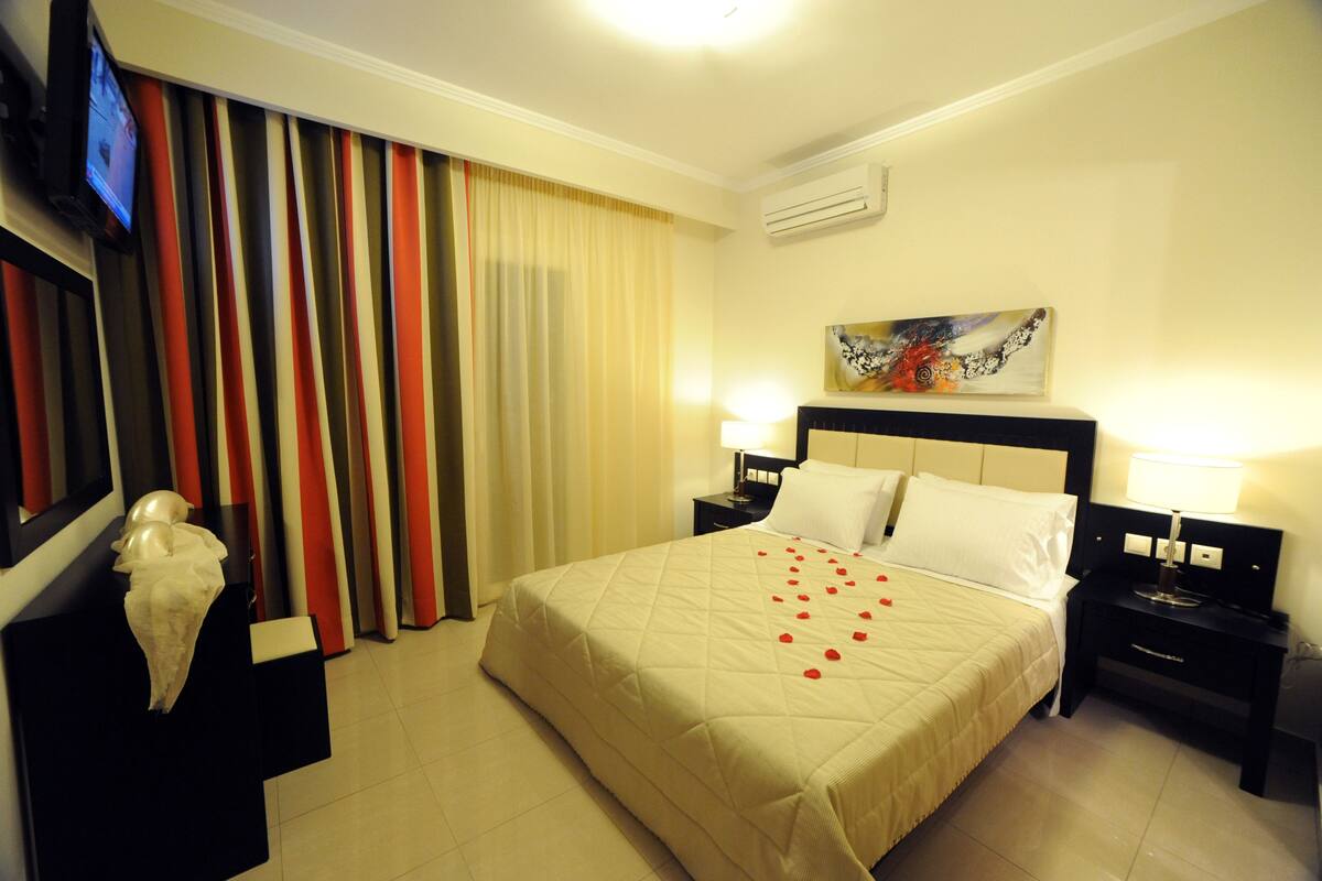 A hotel room with a bed, two bedside tables, a TV, a painting, and a decorative sculpture.