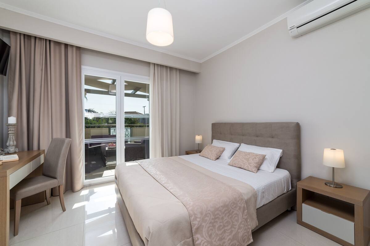 A hotel room with a bed, a desk, a chair, a nightstand, and a large window with curtains.