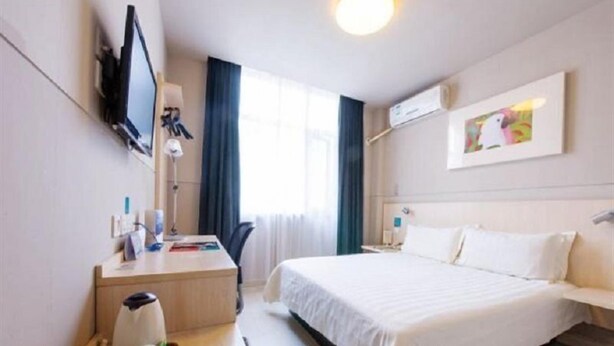 A hotel room with a bed, a desk with a chair, a TV mounted on the wall, and a window with curtains.