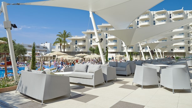 Zona junto a la piscina con mobiliario exterior moderno y sombrillas, un cielo azul despejado y una piscina al fondo.