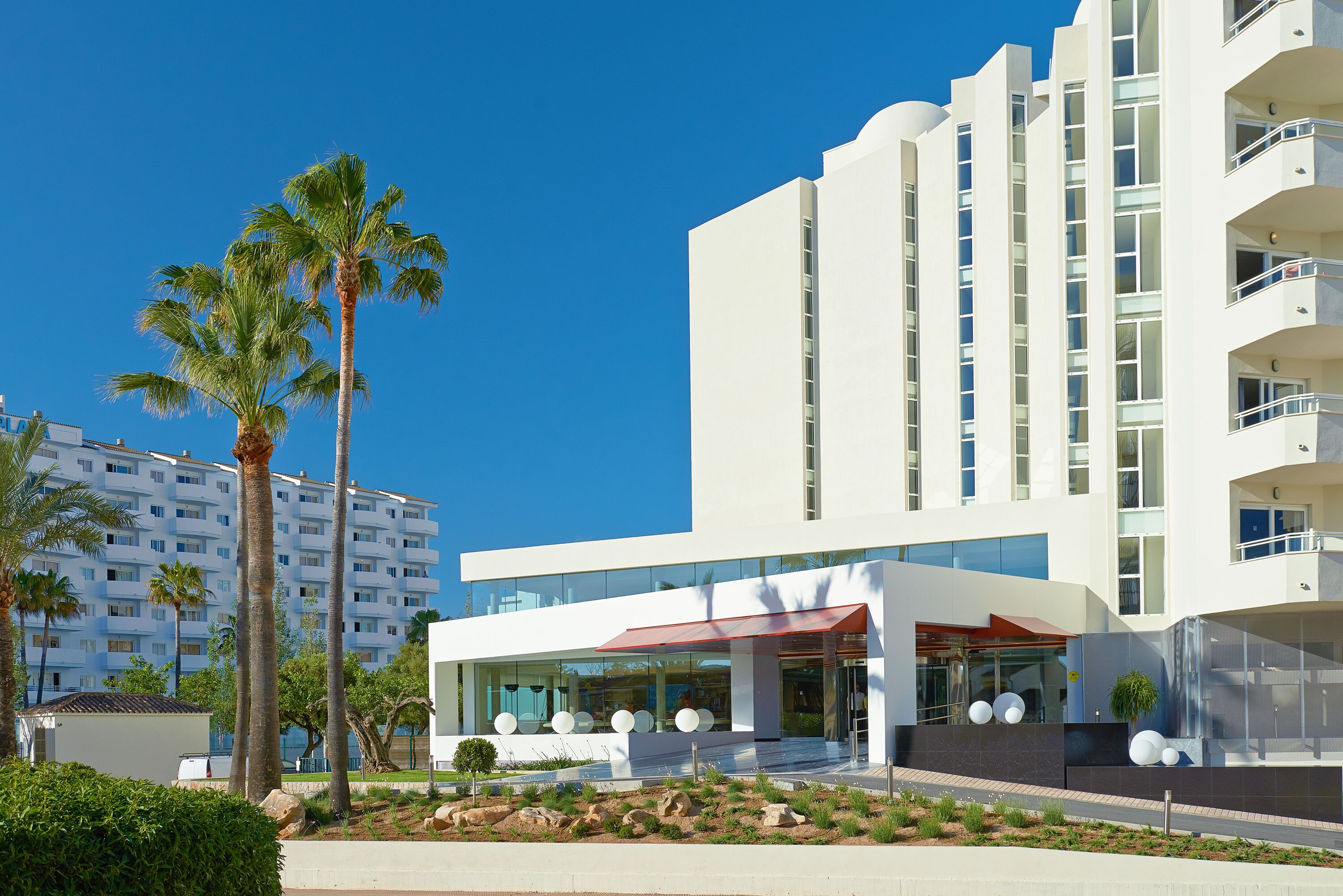 A modern hotel with a white facade, large windows, and a red awning.