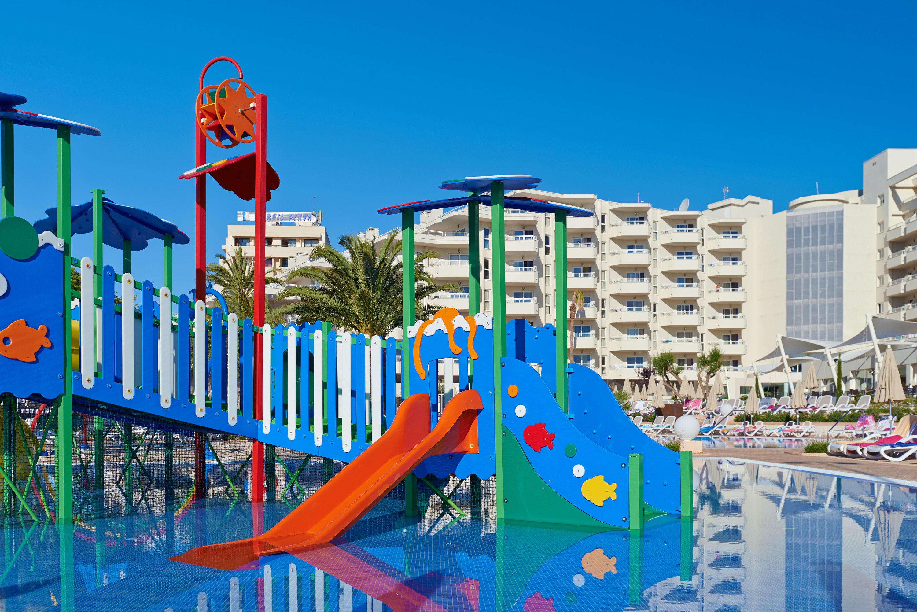 A children's play area with slides and climbing structures, a pool, and a beachfront hotel in the background.