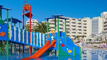 A children's play area with slides and climbing structures, a pool, and a beachfront hotel in the background.