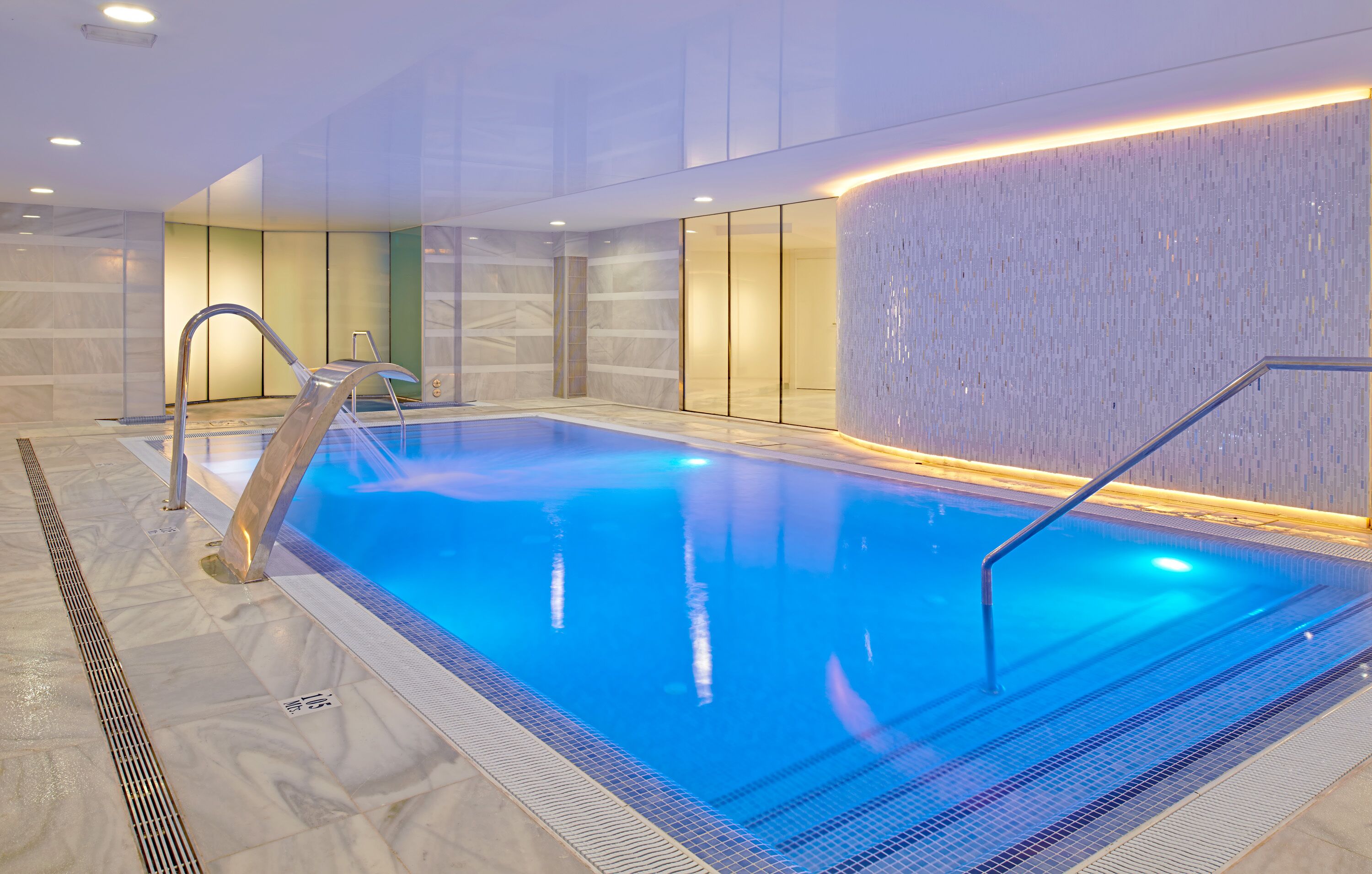 Una piscina interior con agua cristalina, una escalera metálica y suelo de baldosas.