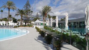 A pool area with lounge chairs and umbrellas, surrounded by palm trees and a modern building. - Canary Garden Club (San Bartolomé de Tirajana)