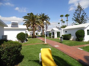 A courtyard with white buildings, palm trees, and a yellow lounge chair. - Canary Garden Club (San Bartolomé de Tirajana)