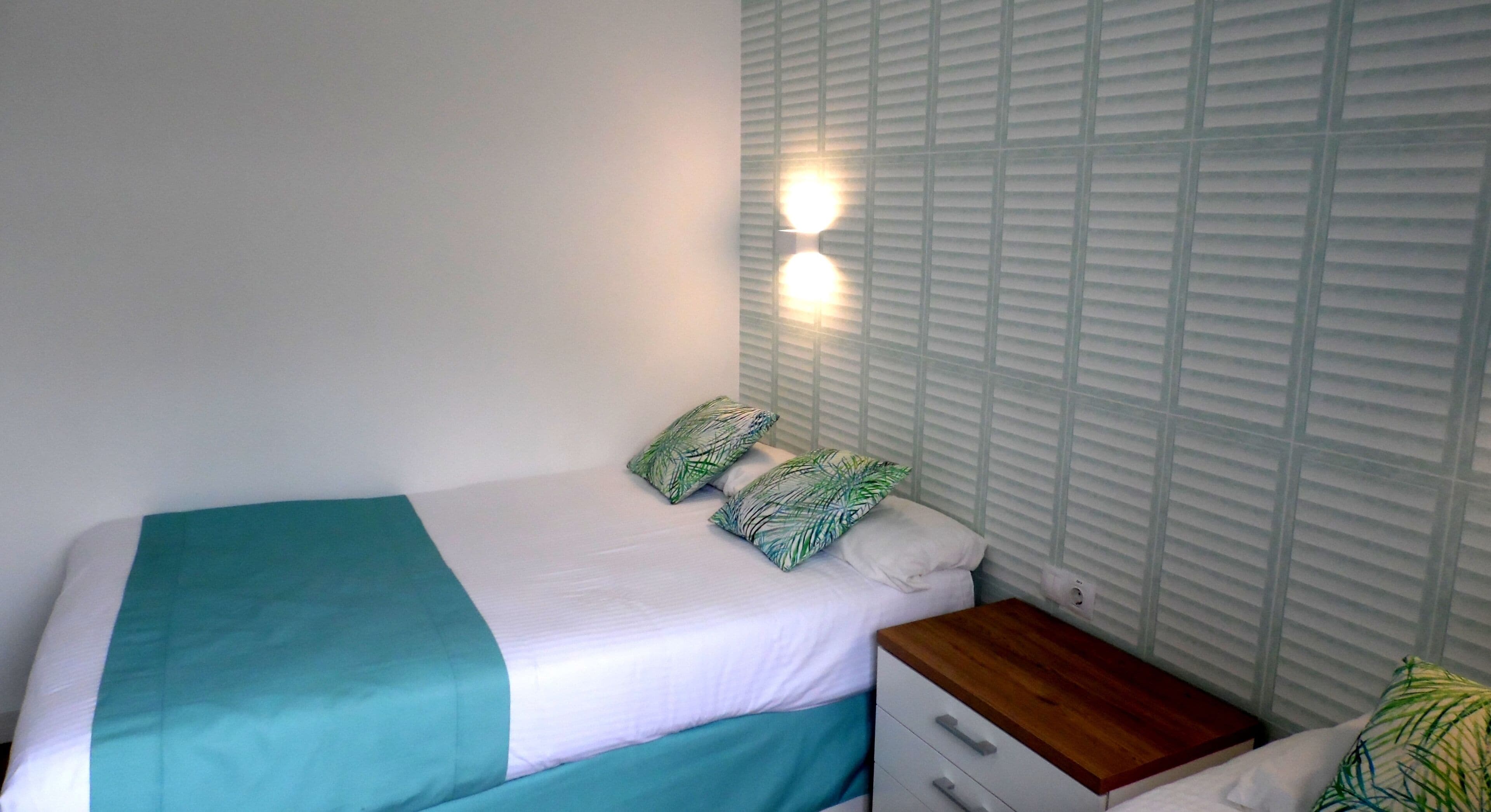 A bedroom with a bed, two pillows, a nightstand, and a wall-mounted light.