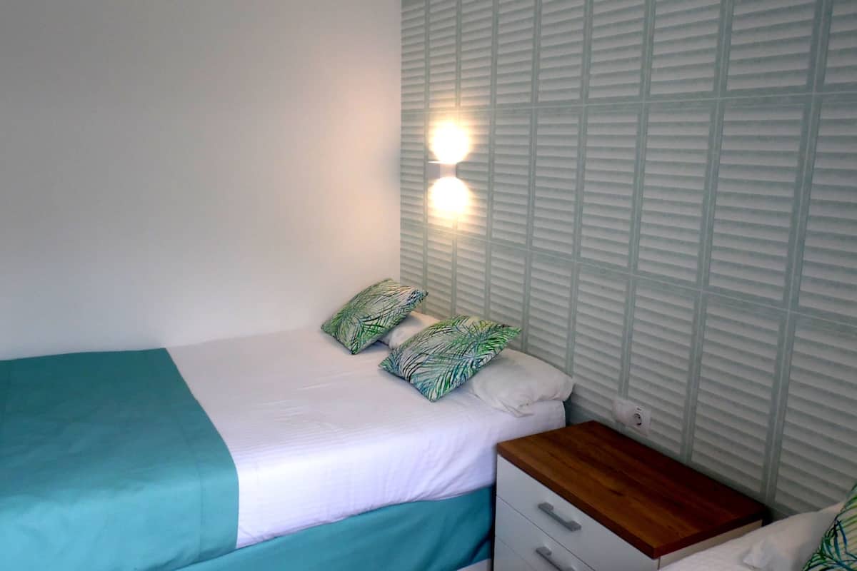 Un dormitorio con una cama, dos almohadas, una mesita de noche y una lámpara fijada a la pared.