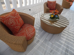 A wicker sofa with a red patterned cushion, a round glass coffee table with a floral centerpiece, and a white outdoor area with a pebble path. - Canary Garden Club (San Bartolomé de Tirajana)