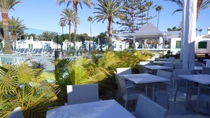 A poolside area with white tables and chairs, surrounded by palm trees and a clear sky. - Canary Garden Club (San Bartolomé de Tirajana)
