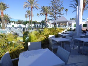 A poolside area with white tables and chairs, surrounded by palm trees and a clear sky. - Canary Garden Club (San Bartolomé de Tirajana)