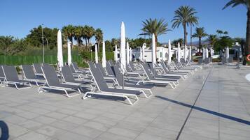 Een rij lege ligbedden met parasols, gerangschikt op een betegeld terras met palmbomen op de achtergrond.