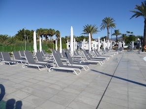 Eine Reihe leerer Liegestühle mit Sonnenschirmen, angeordnet auf einer gefliesten Terrasse, im Hintergrund Palmen.