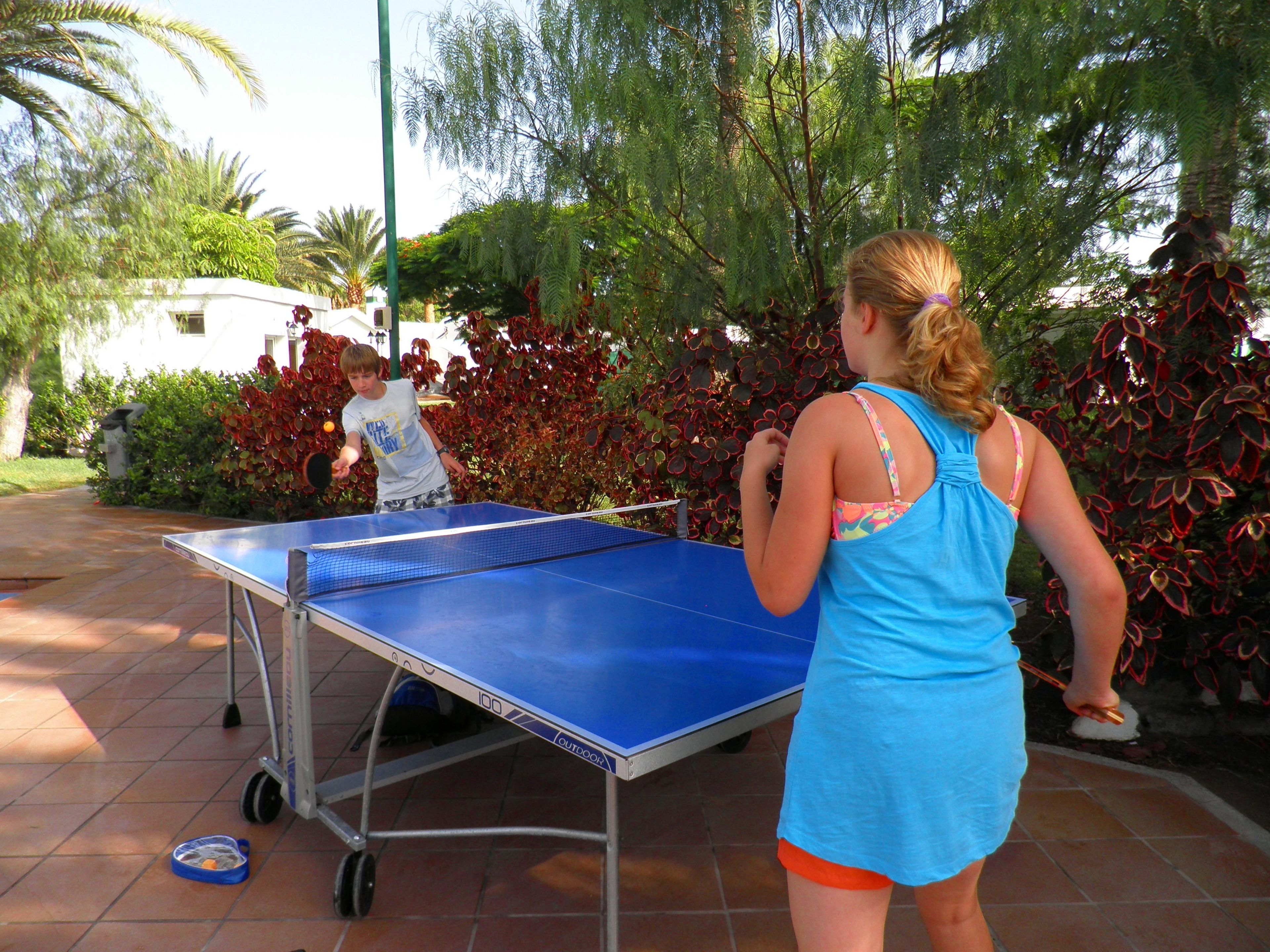 Mesa de ping pong al aire libre con estructura metálica y superficie de juego azul, rodeada de follaje y piso de baldosas.
