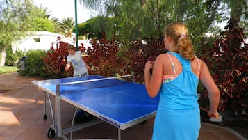 Mesa de ping pong al aire libre con estructura metálica y superficie de juego azul, rodeada de follaje y piso de baldosas.